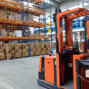 WMS es una forma de almacenamiento, la imagen es de un montacargas naranja, en donde hay varios estantes llenos de cajas.